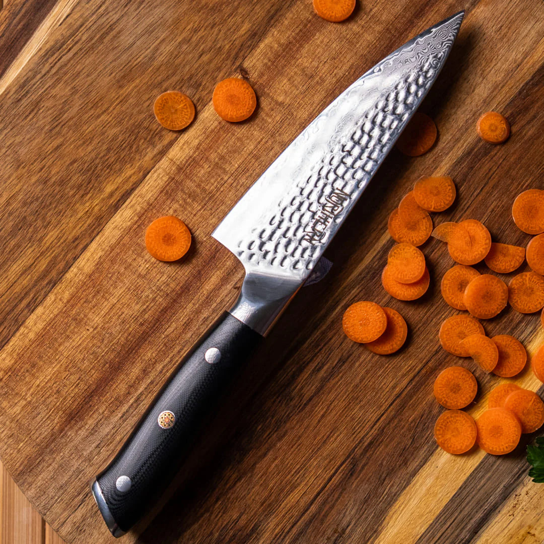 Northern Knife Odin Chef Knife with hammered Damascus steel blade resting on a wooden cutting board next to freshly sliced carrots, highlighting its tsuchime finish and precision edge.