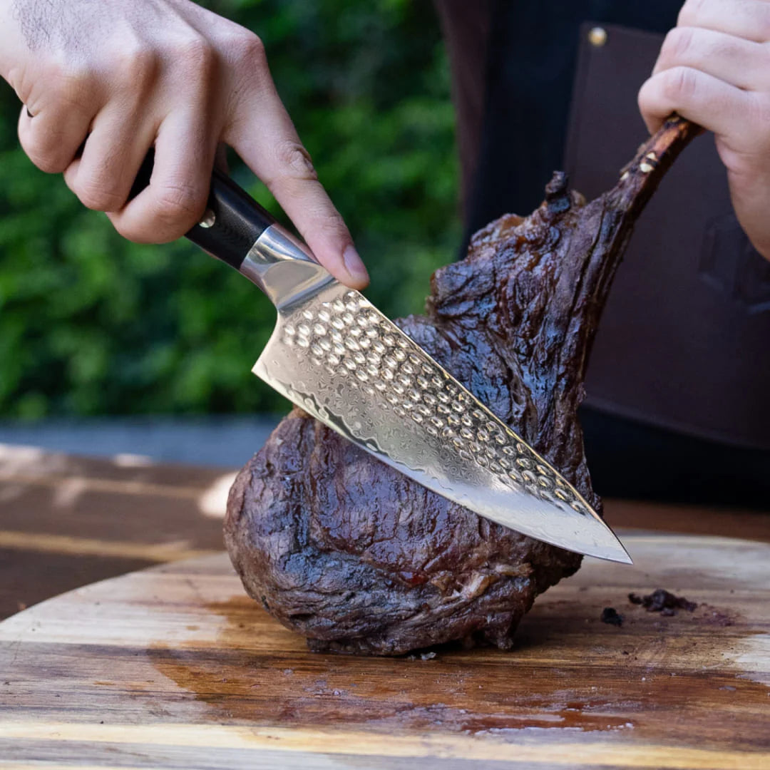 Northern Knife Odin Chef Knife with hammered tsuchime finish slicing a large tomahawk steak on a wooden carving board.